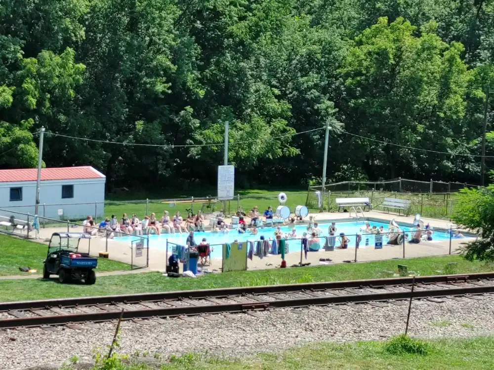 Pool with people swimming, sitting on the side, and a small building next to it, near a train track.