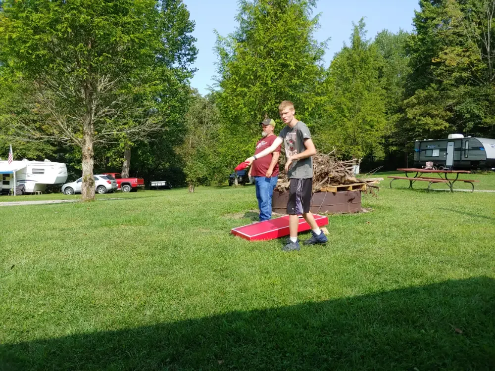 Two people playing cornhole in a grassy campground, with RVs and trees in the background.