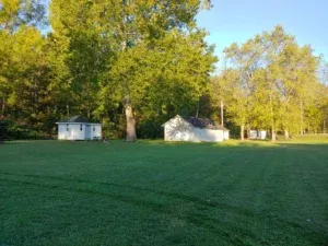 Green lawn with two small white buildings and trees in the background, under a blue sky.