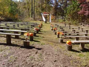 Outdoor wedding ceremony setup with benches, pumpkins, and an arch in a wooded area.