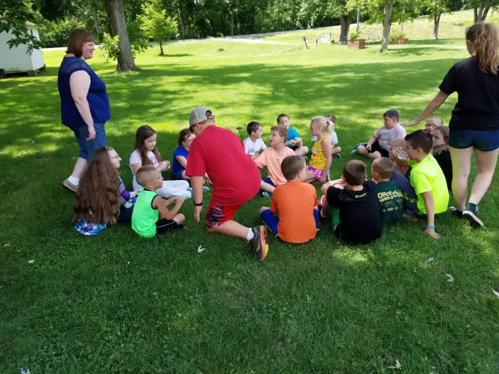 Group of children sitting in a circle on grass, with two adults. The children are looking at the person in the middle.