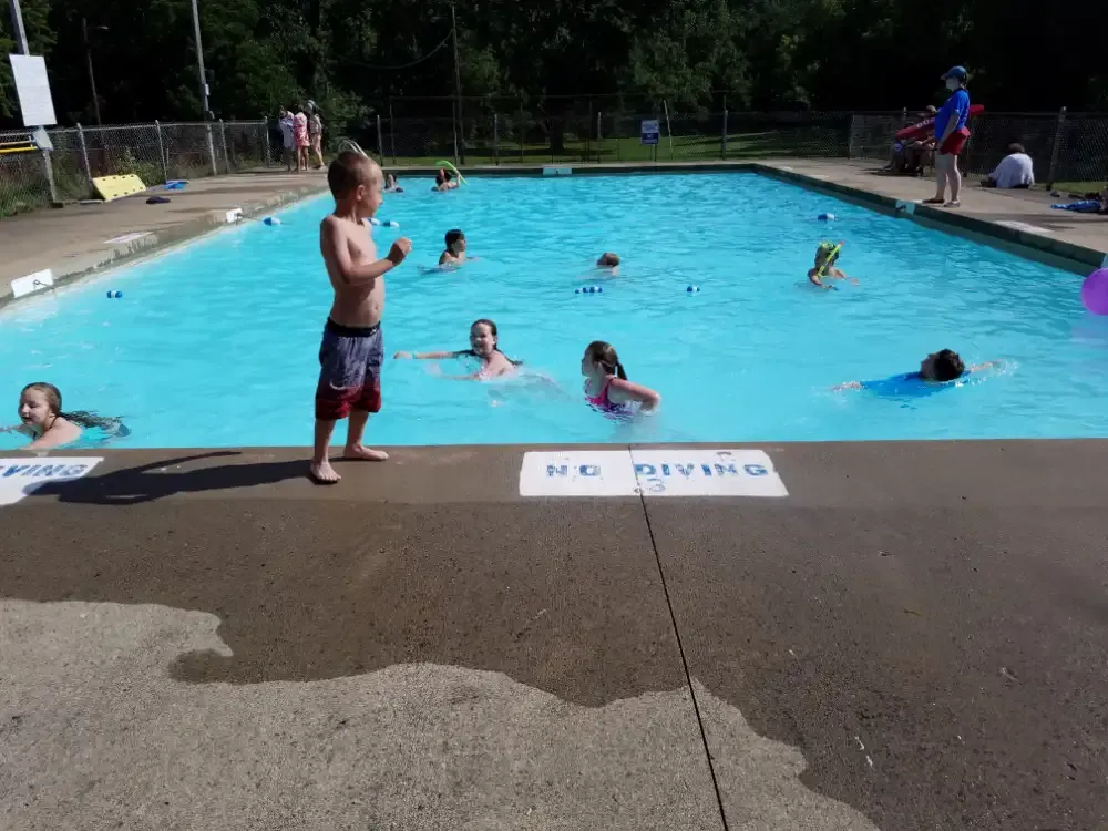 Children swimming in a blue pool on a sunny day; a boy stands by the edge.