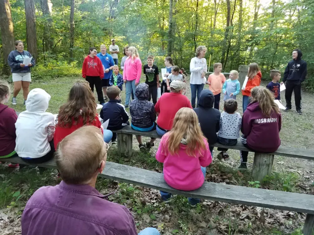 Group of children and adults in a forest, gathered around benches. They appear to be listening.