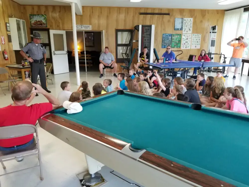 A group of kids listens to a police officer in a rec room with a pool table and ping pong table.