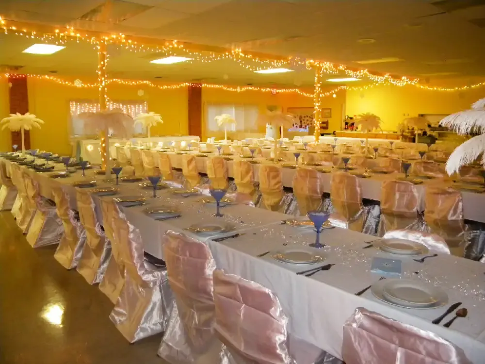 A banquet hall with long tables set for a formal event. Tables have white linens, pink chair covers, and blue glasses.
