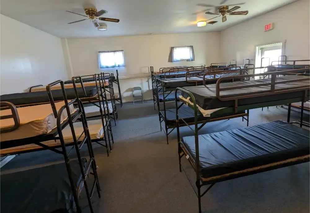 Inside a dorm room, filled with rows of bunk beds, a door and windows are seen.