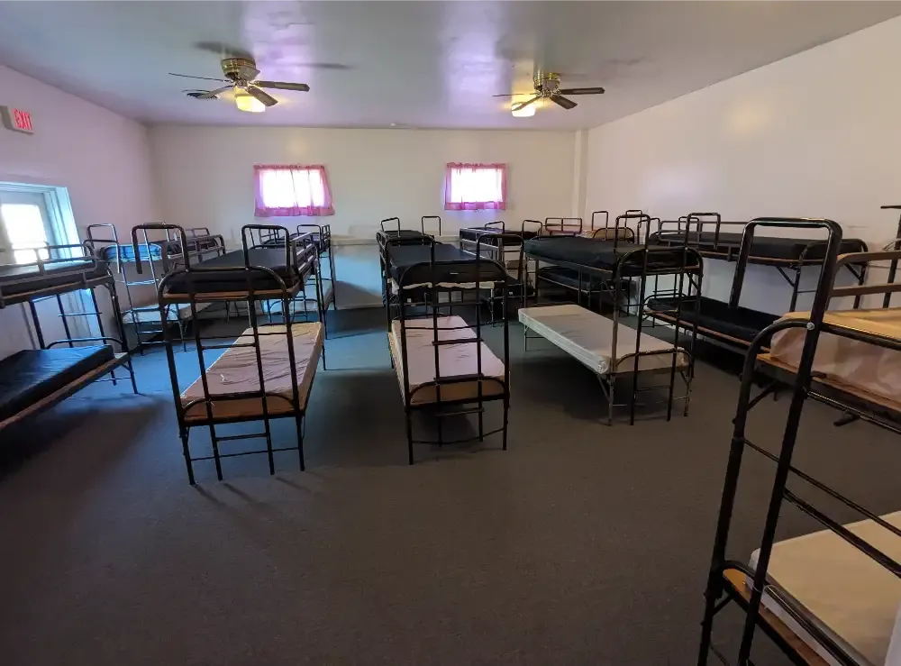 Inside view of a large room with multiple bunk beds. White walls, ceiling fans, and windows with pink curtains.