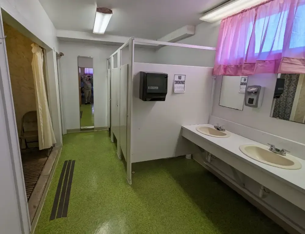 Public restroom with green floor, white walls, sinks, and pink curtains on a window.