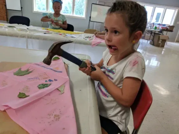 Girl with shocked expression holding a hammer above pink fabric at a craft table.