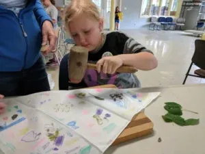 Young person uses a mallet to work on a piece of fabric with drawings on a table.