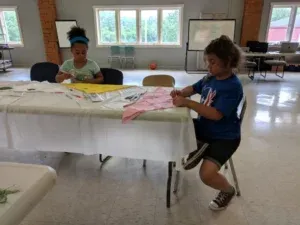 Two children crafting at a table in a bright room. One is kneeling, another seated. Clothes are on the table.