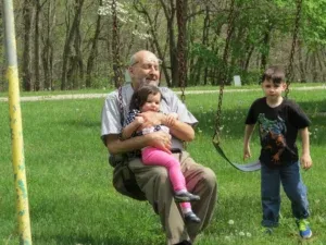 An elderly man swings with a young child on his lap, a boy stands nearby on a grassy field.