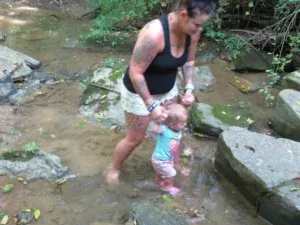 Woman with tattoos helps a baby walk in a shallow creek, surrounded by rocks and greenery.