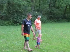 Man, woman, and toddler walking in a grassy field. Man holds toddler's hand.