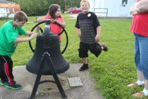 Children playing with a large black bell in a grassy park. One boy pulls a wheel, another kicks in excitement.