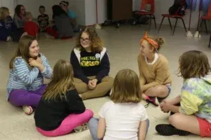 Group of girls sitting in a circle, talking. They're in a room with other people in the background.