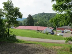 A long, white building with a red roof in a grassy area, with trees and a hill in the background.