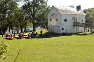 White building, people sitting on the grass, trees and water in the background.