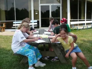 Children at a picnic table writing outside a building. One girl smiles at the camera.