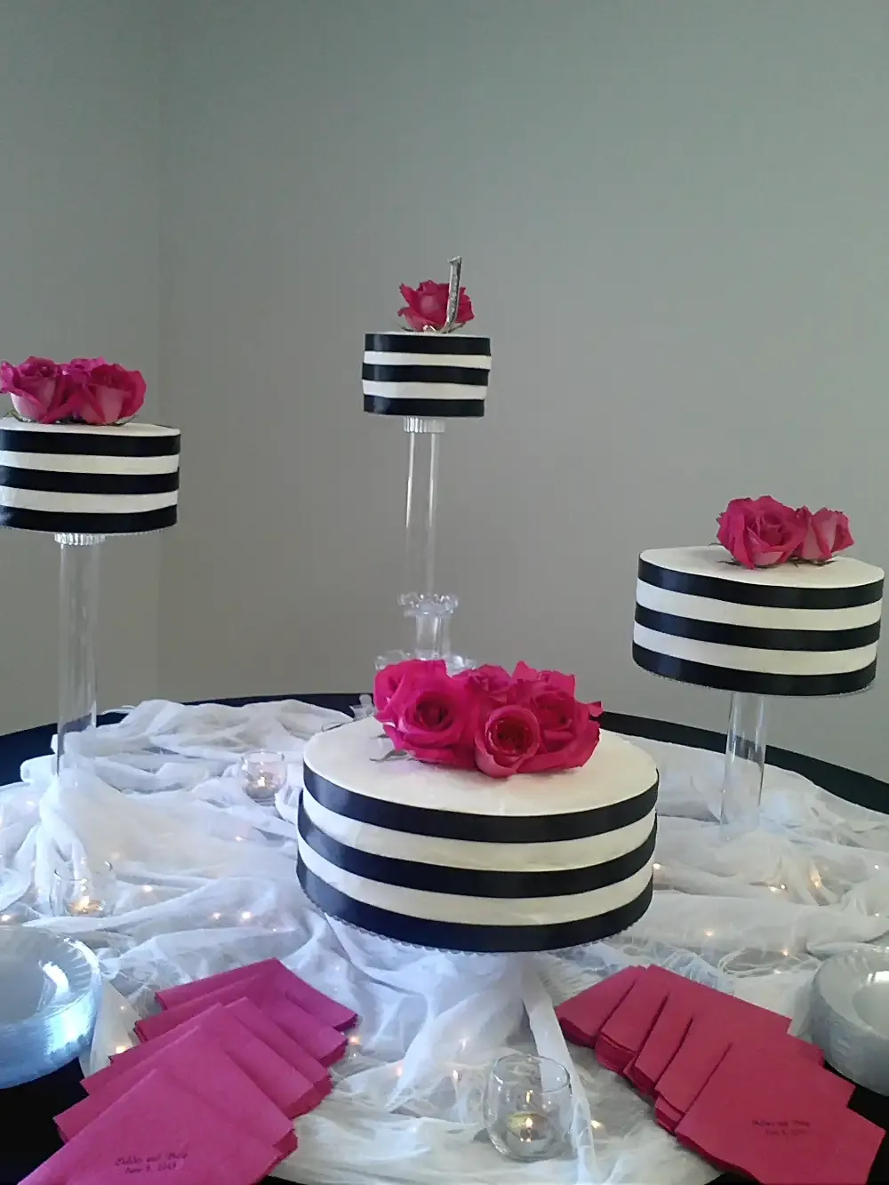 Black and white striped cakes with pink roses, set on clear stands, arranged on a table with white fabric and pink napkins.