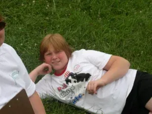 A young person with reddish-blonde hair wearing a white t-shirt reclines on green grass, smiling.