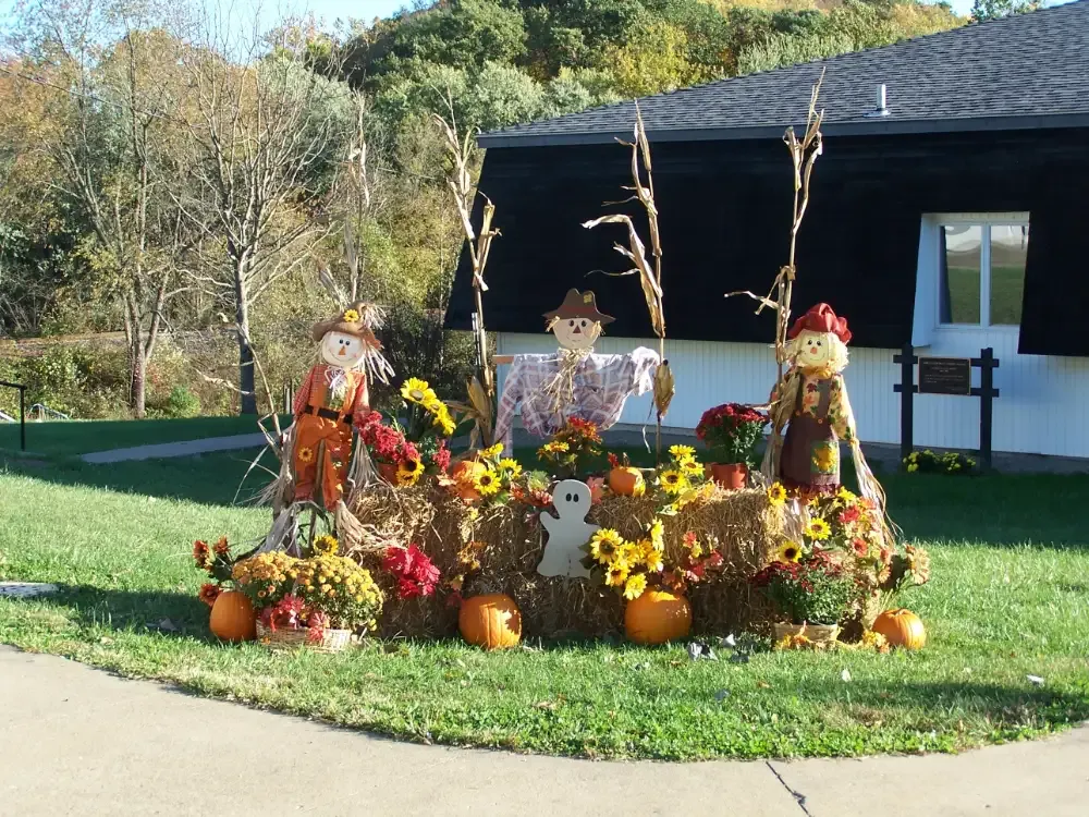 Fall display: scarecrows, hay bales, pumpkins, mums, and corn stalks on a lawn in front of a building.