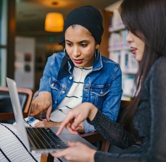 Two women collaborating on a laptop in a café, pointing at the screen.