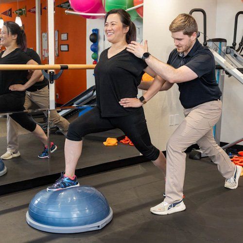 Two people exercising in a gym, balancing on a BOSU ball during a guided workout.