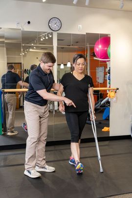 Trainer assists person walking with crutches in a gym studio mirror room.
