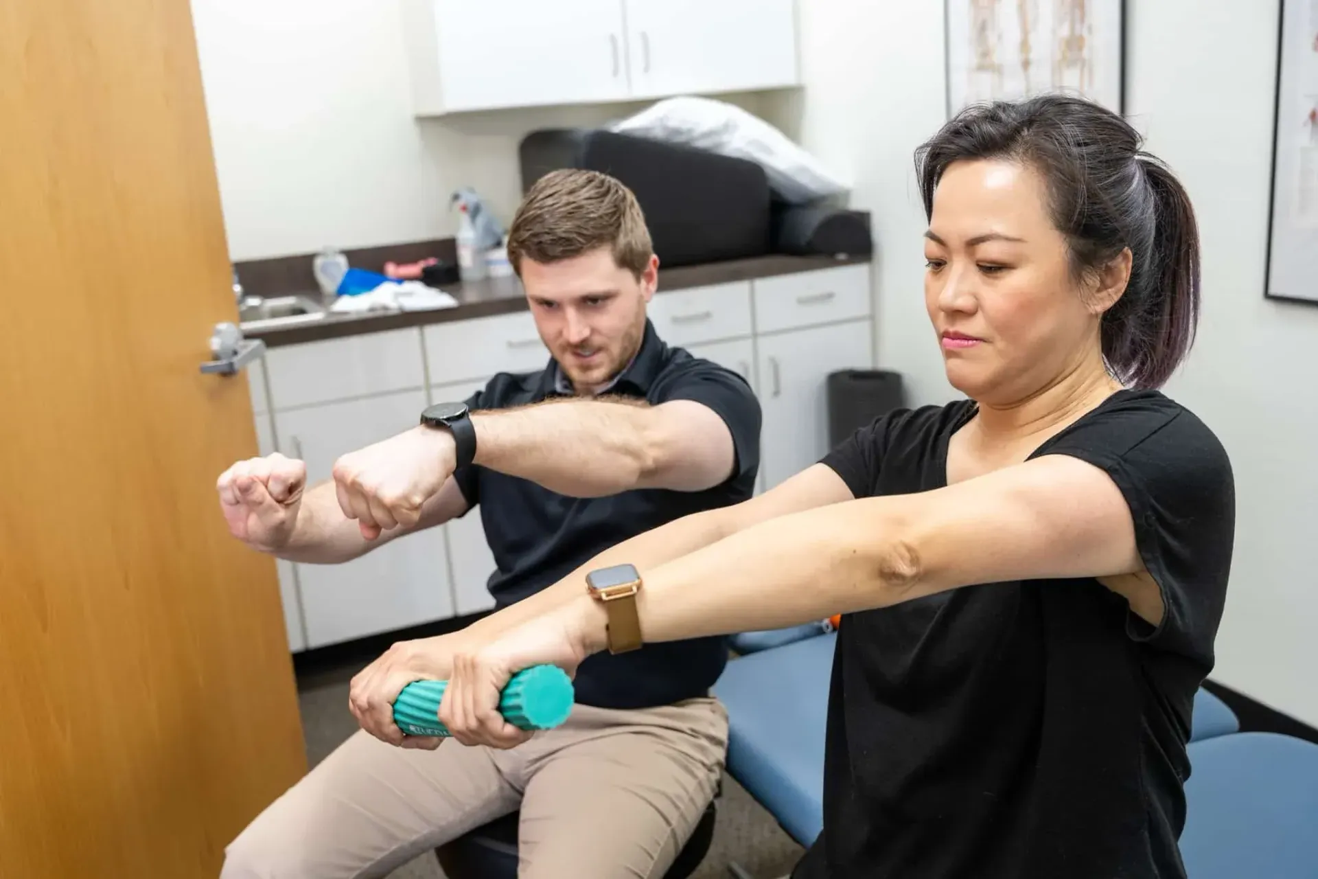 Two people extending their arms in a clinical exercise room, wearing wrist weights or trackers.