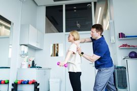 Two people in a bright kitchen, one helping the other stretch a shoulder.