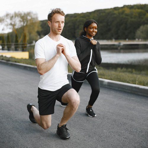 Two people doing lunges on a road beside water, wearing athletic clothes.