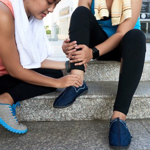 Person holding an ankle on outdoor steps, suggesting a foot injury while another person kneels beside them.