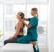 Physical therapist in teal scrubs treating a seated patient on a white exam table by a bright window