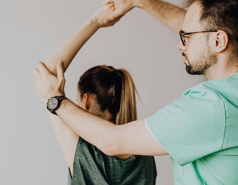 Two people stretching arms overhead in a bright room; one guides the other’s pose.
