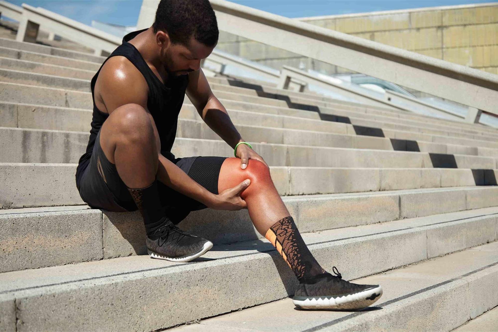 Athlete seated on outdoor steps, holding and rubbing his injured knee with a red brace.