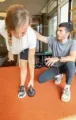 Person assisting another with balance exercise in a gym, on orange floor near mirrors