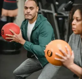 People doing medicine ball exercises in a gym, one holding a red ball and another an orange ball.