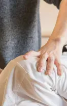 Hands massage a person’s shoulder in a close-up, with a gray shirt and white fabric visible.