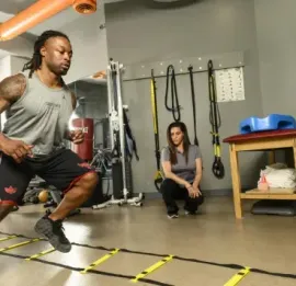 Two people training in a gym, one doing agility drills beside yellow ladder markers.