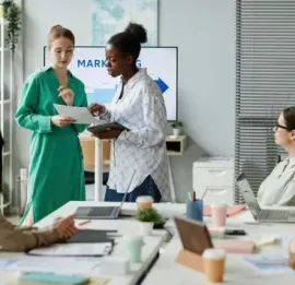 Two coworkers discussing papers in a bright office meeting room with laptops and coffee cups.
