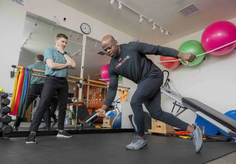 Two people doing a resistance band workout in a gym, with exercise balls and equipment in the background.