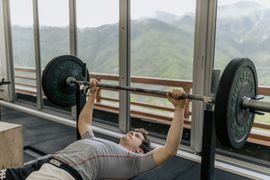 Person bench pressing a barbell in a gym with large windows and mountain views.