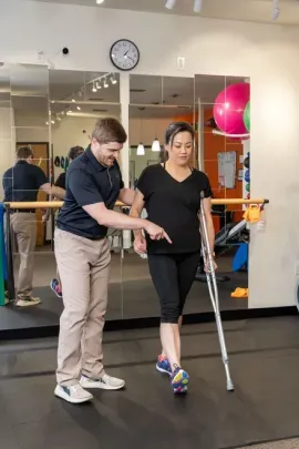 Trainer assists person walking with crutches in a gym studio mirror room.