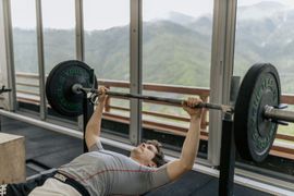Person bench pressing a barbell in a gym with large windows and mountain views.