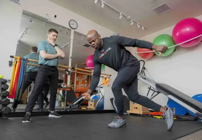 Two people doing a resistance band workout in a gym, with exercise balls and equipment in the background.