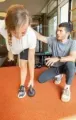 Person assisting another with balance exercise in a gym, on orange floor near mirrors