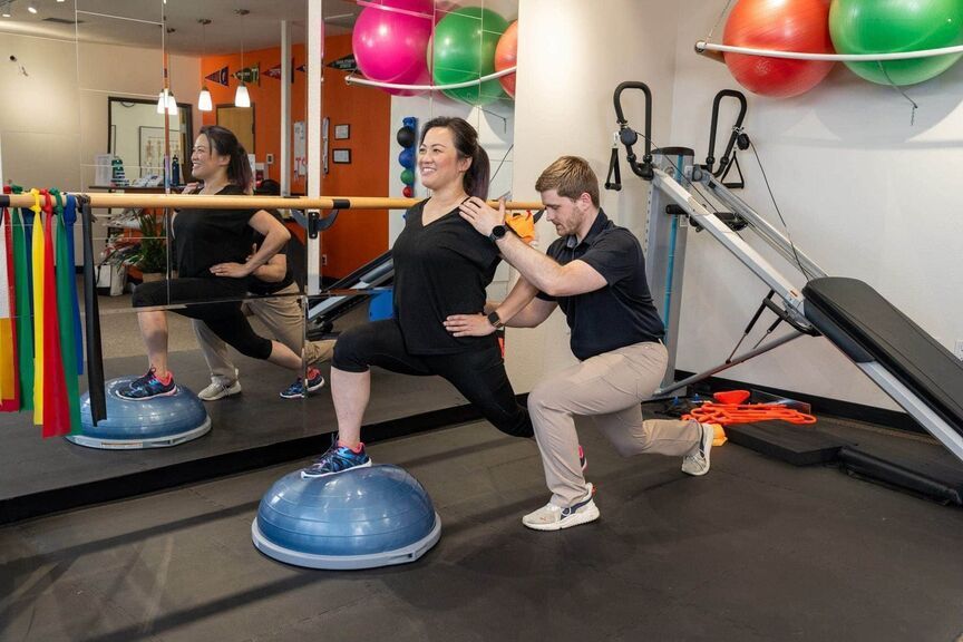 Two women exercising in a gym, doing lunges and balancing on a blue BOSU ball with a trainer assisting.
