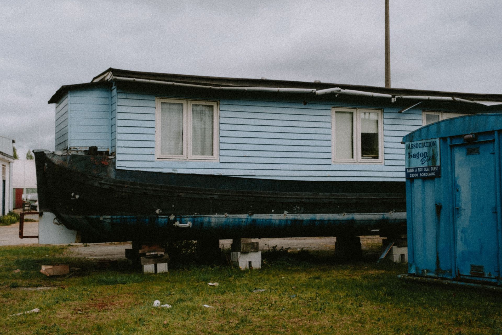 Blue mobile home elevated on blocks beside a blue shed in a grassy yard