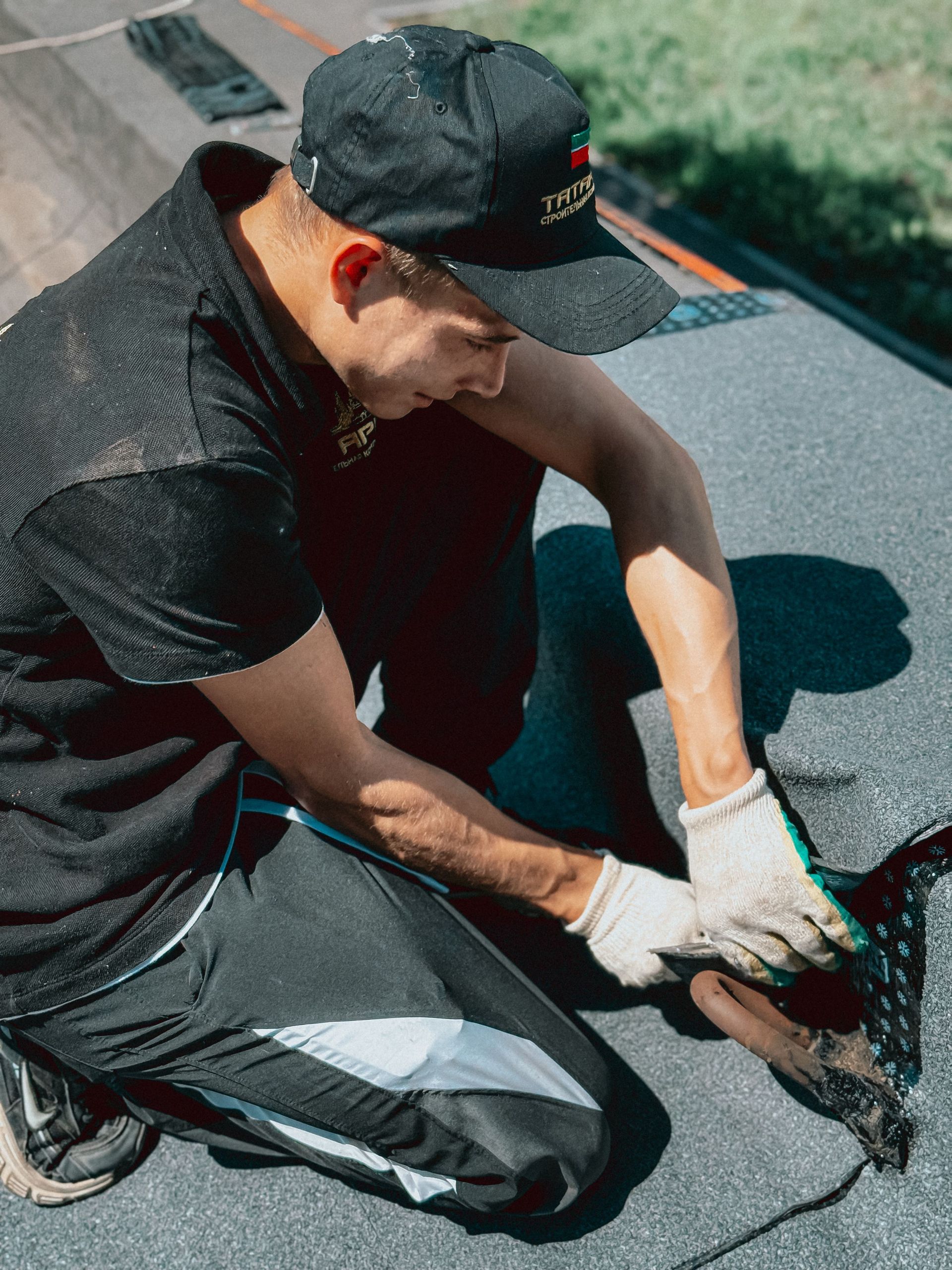 Person in a black shirt and cap wearing gloves, kneeling on a roof using specialized tools for repair.